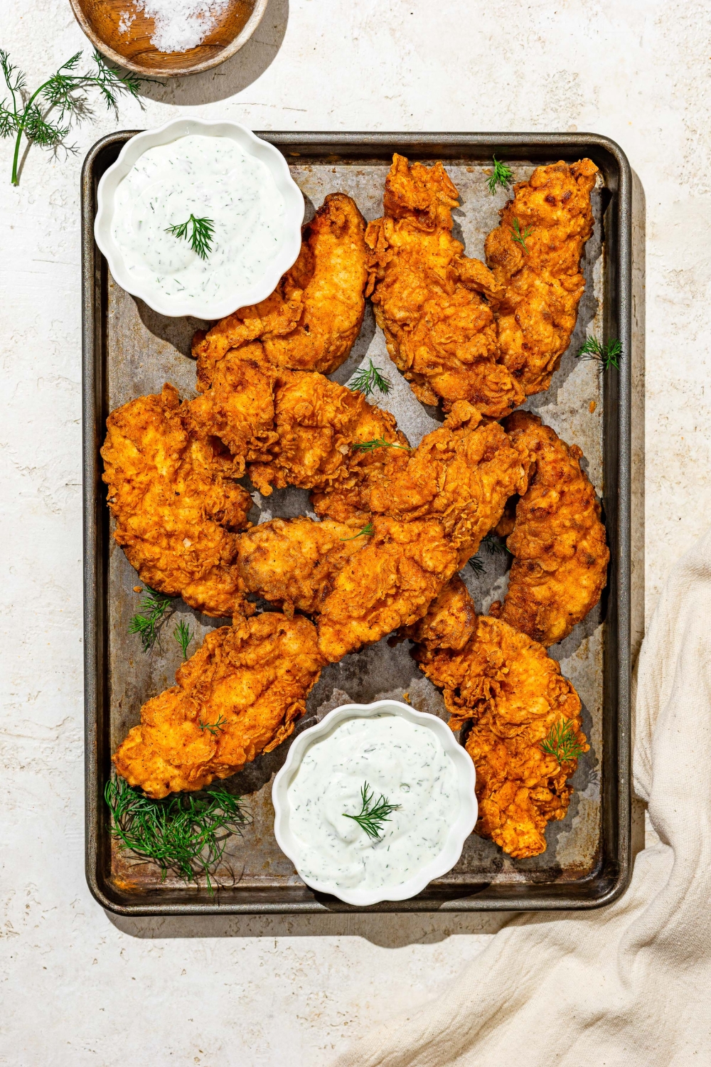 A baking sheet with buttermilk fried chicken tenders with two bowls of homemade ranch dressing. The sheet is on a white counter and garnished with sprigs of dill.