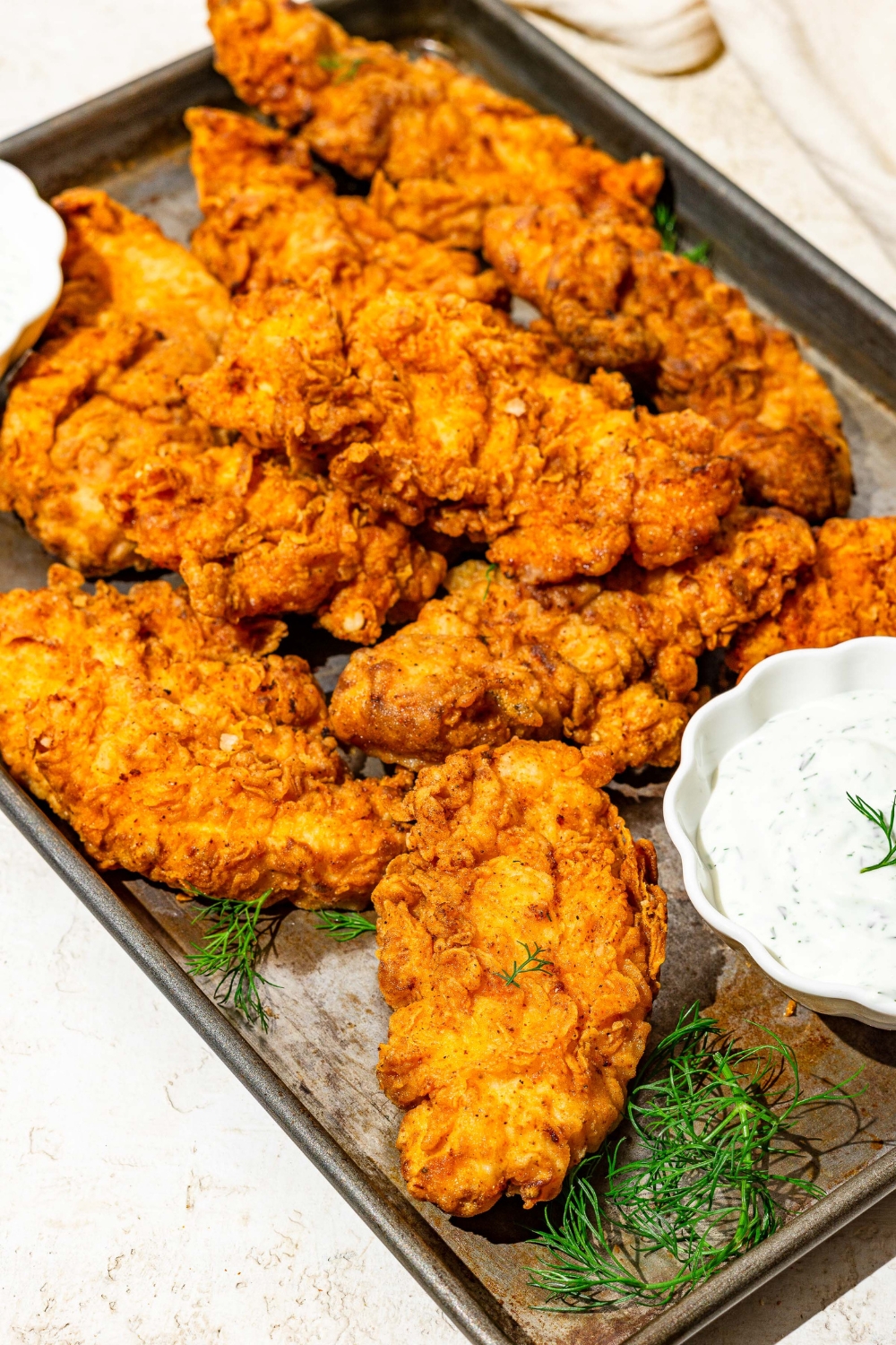 A baking sheet with buttermilk fried chicken tenders with two bowls of homemade ranch dressing. The sheet is on a white counter with a white cloth napkin and garnished with sprigs of dill.