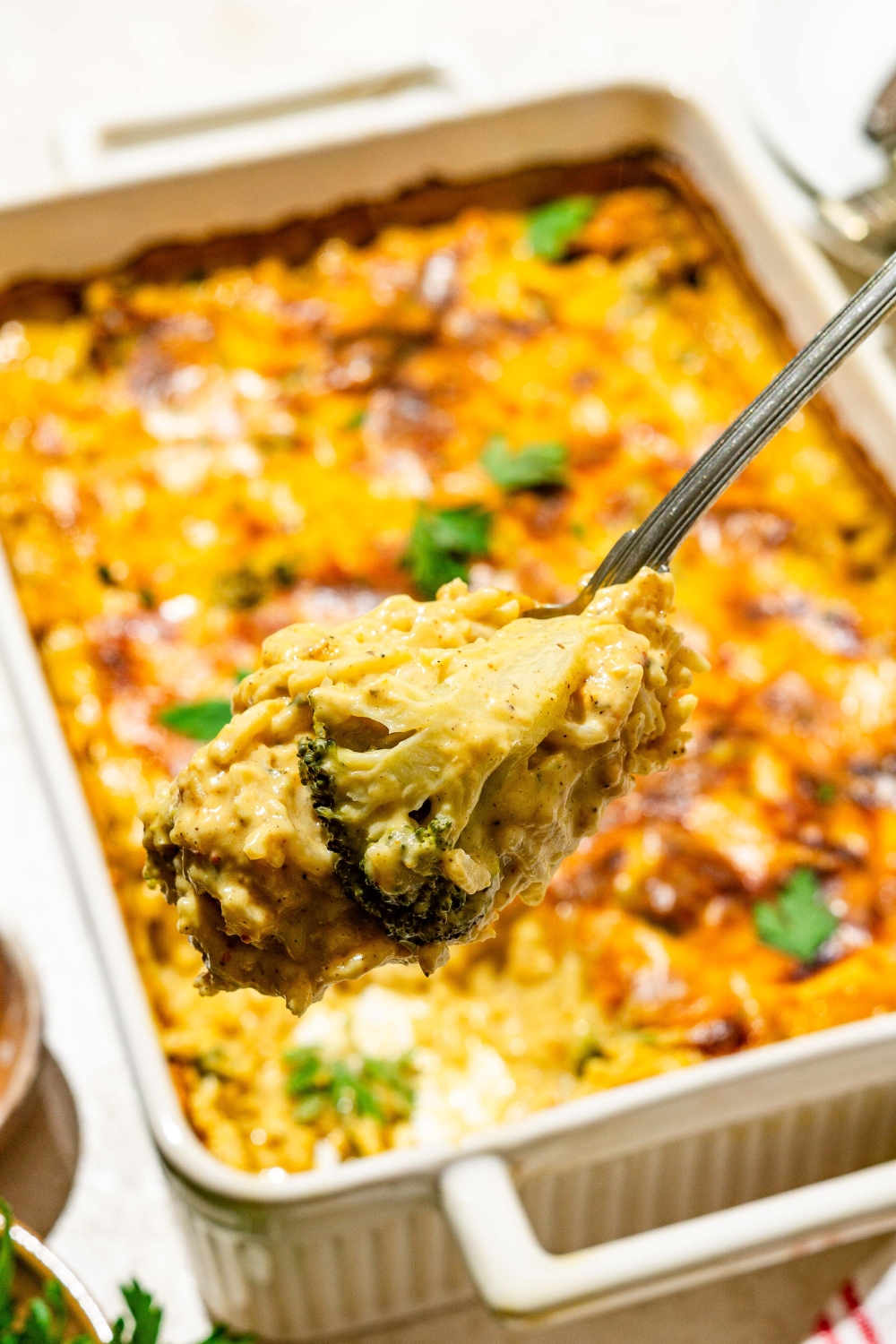 A close up of a spoon with a bite of chicken broccoli rice casserole. There is a baking dish of casserole garnished with fresh parsley blurred in the background.