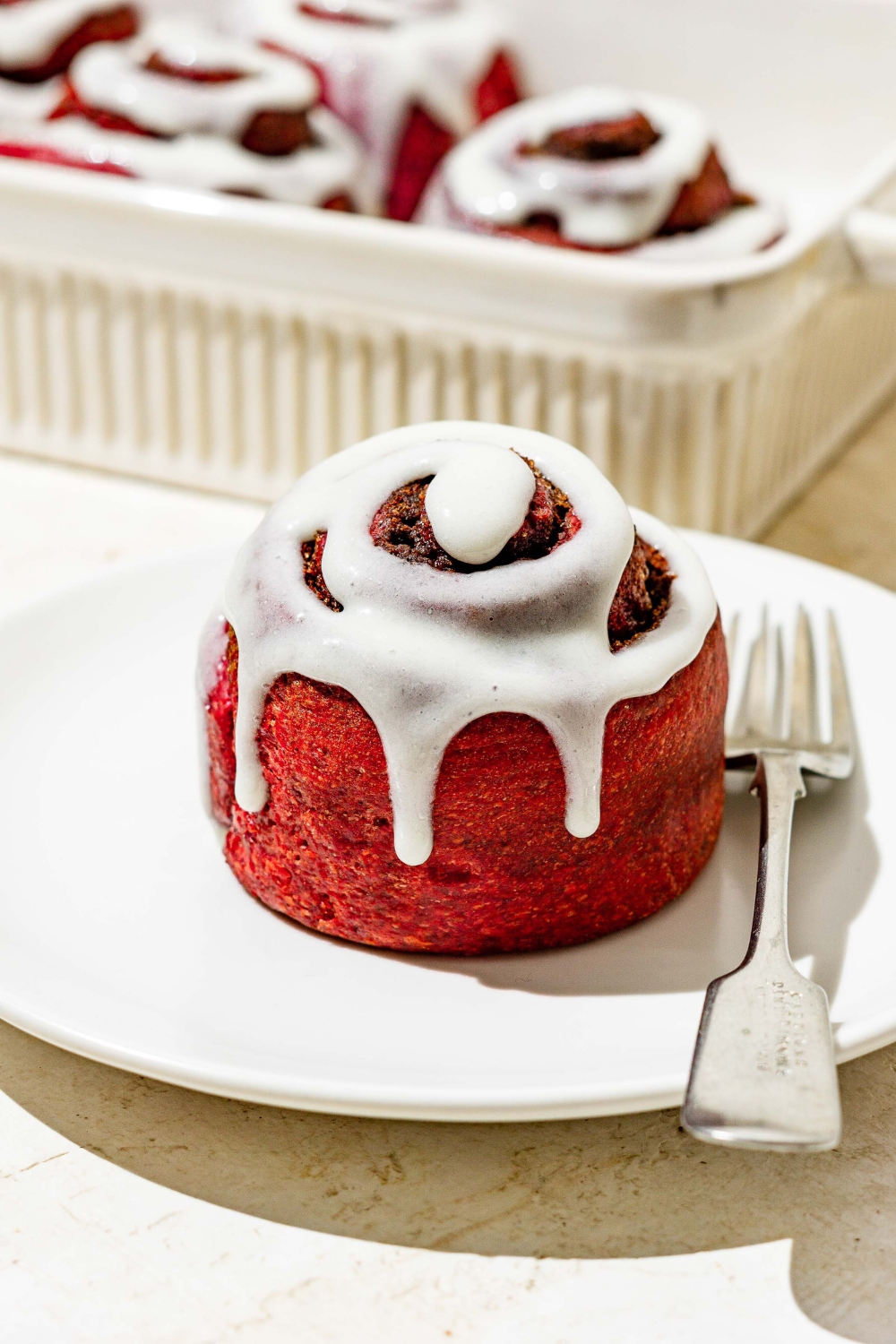 A red velvet cinnamon rolls with cream cheese frosting on a white plate. A baking dish filled with the cinnamon rolls is behind it.