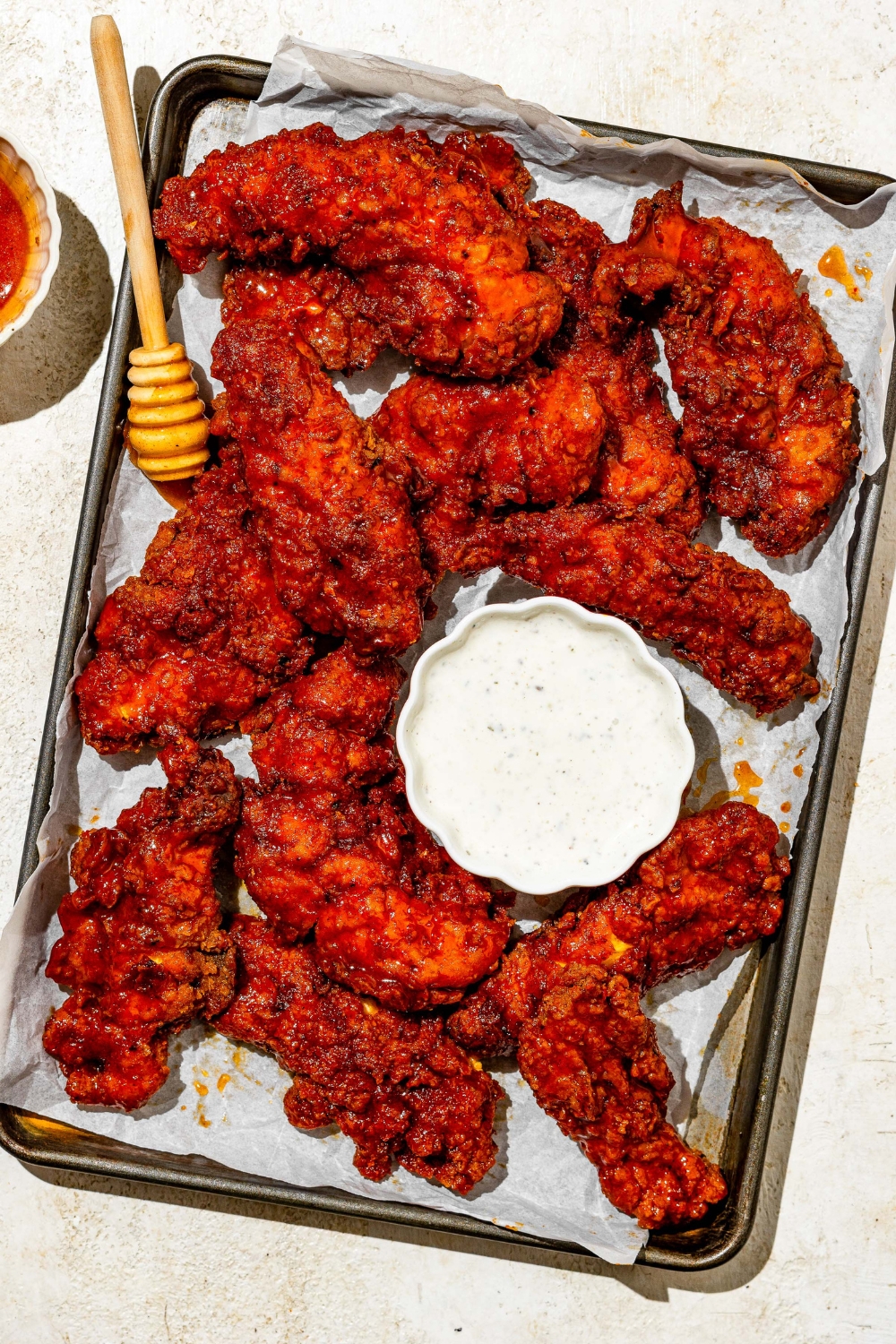A baking sheet lined with parchment paper with copycat Wingstop hot honey chicken tenders. There is a small bowl of ranch sauce and honey stirrer on the baking sheet. The sheet is on a tan counter with a small bowl of honey.