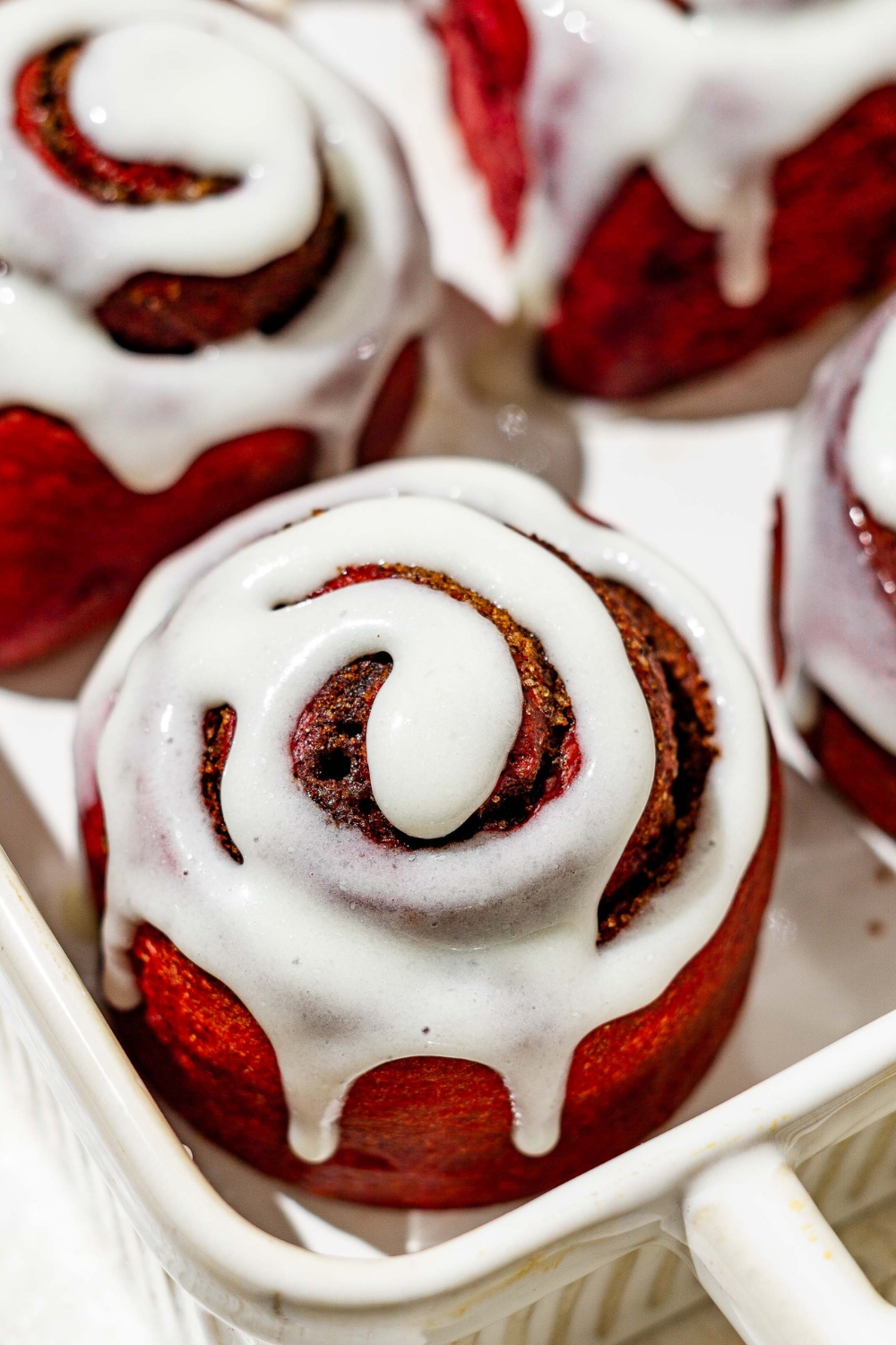 A red velvet cinnamon roll in a baking dish with other red velvet cinnamon rolls around it.