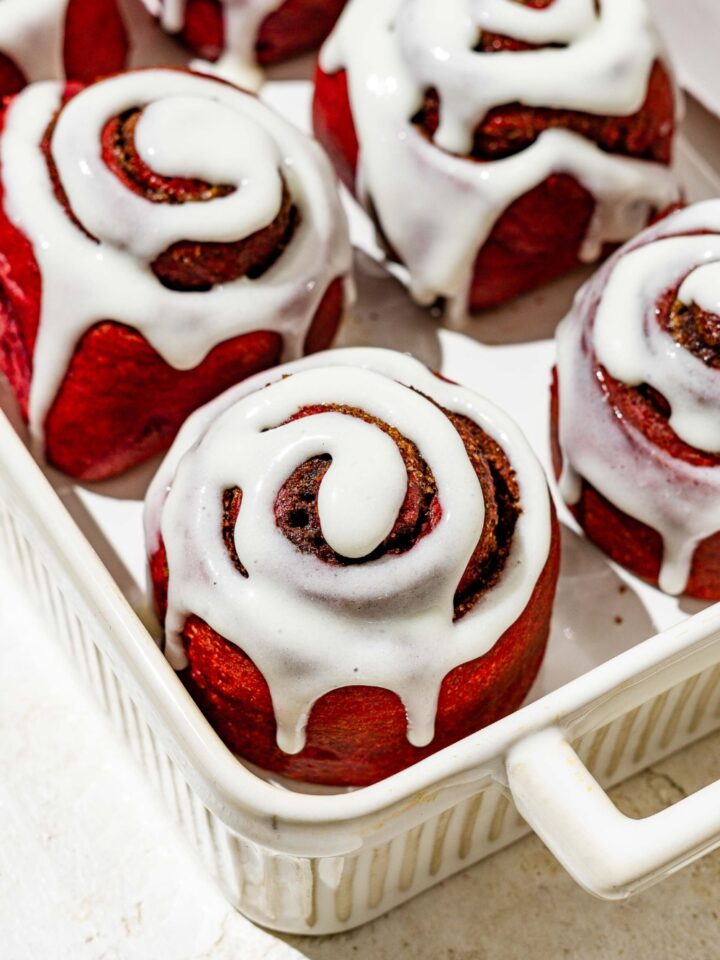 A white baking dish filled with red velvet cinnamon rolls with cream cheese frosting.