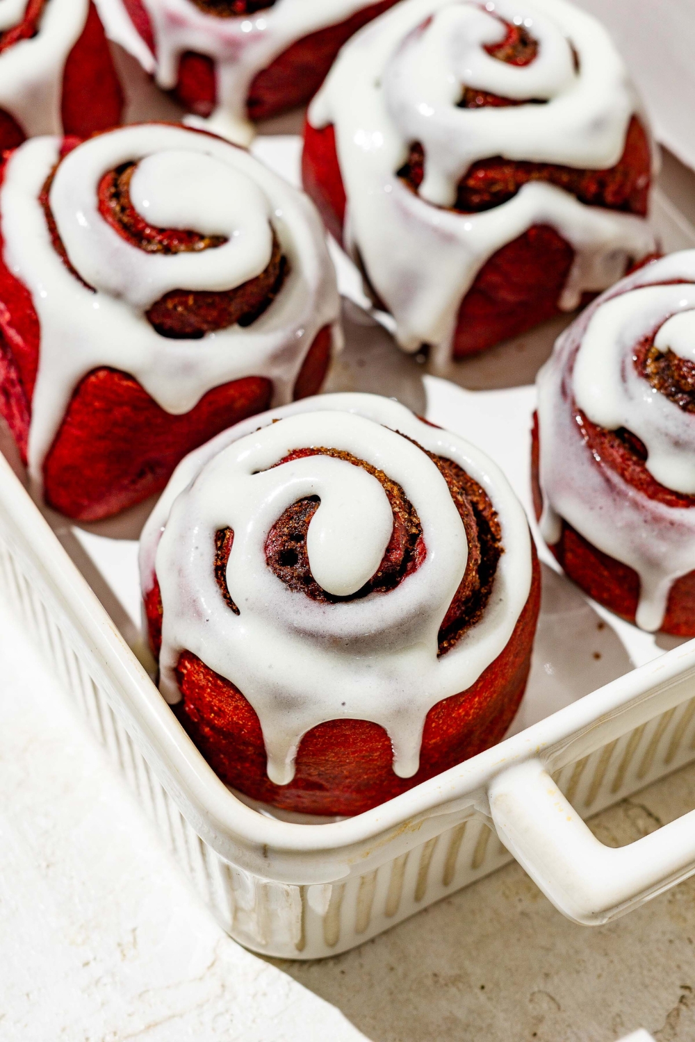 A white baking dish filled with red velvet cinnamon rolls with cream cheese frosting.