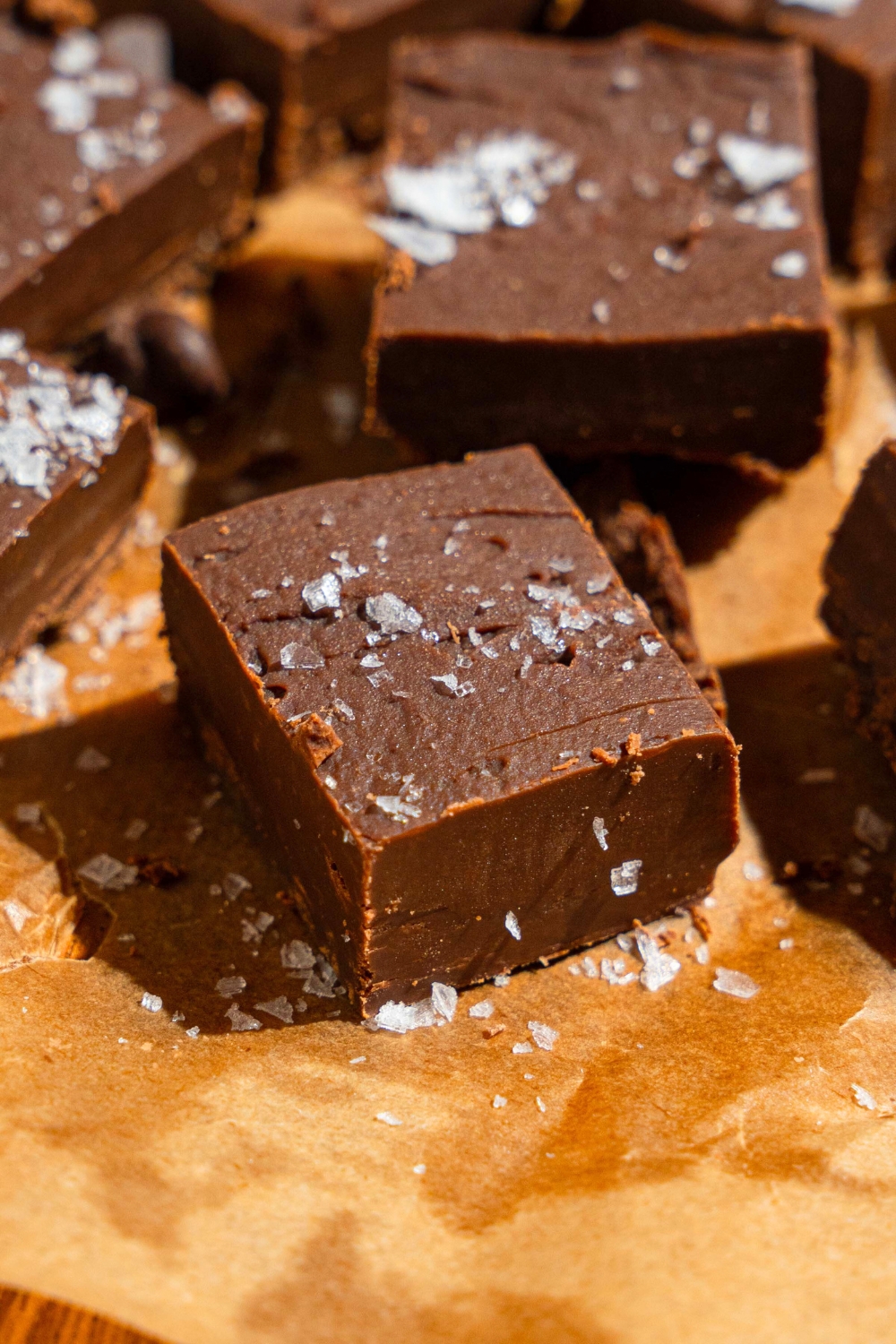 A square of chocolate sweetened condensed milk fudge with flaky salt on top on a sheet of parchment paper with more squares of fudge behind it.