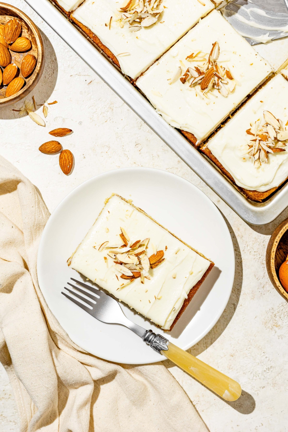 A white plate with a slice of white Texas sheet cat topped with sliced almonds. There is a fork on the plate. The plate is on a white counter with a baking pan with cake and white cloth napkin.