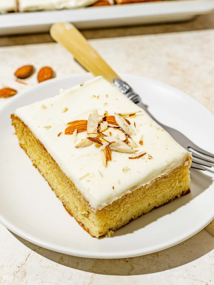 A white plate with a slice of white Texas sheet cat topped with sliced almonds. There is a fork on the plate. The plate is on a white counter with a baking pan with cake.