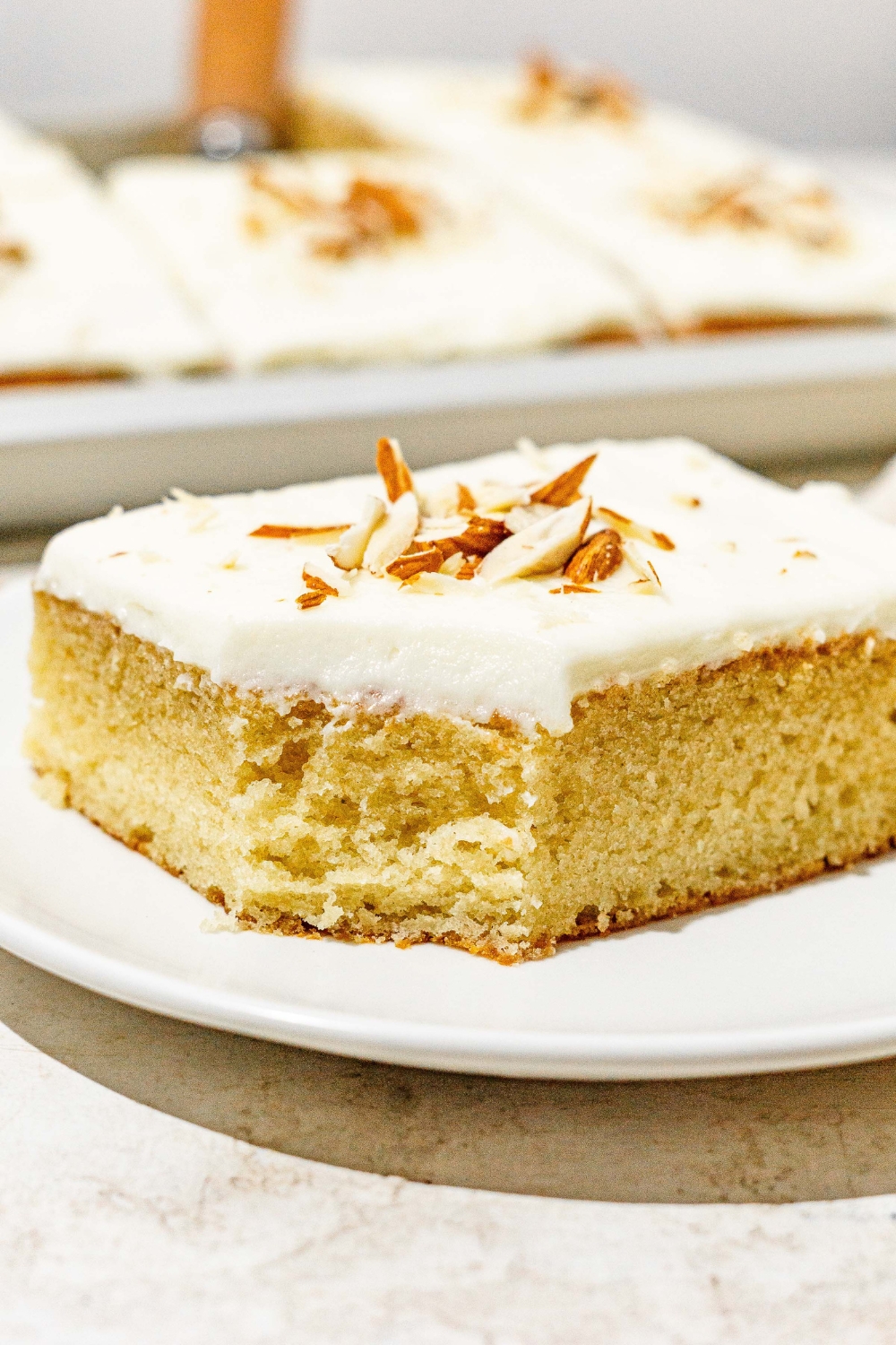 A white plate with a slice of white Texas sheet cat topped with sliced almonds. A bite is taken from the cake. The plate is on a white counter with a baking pan with cake.