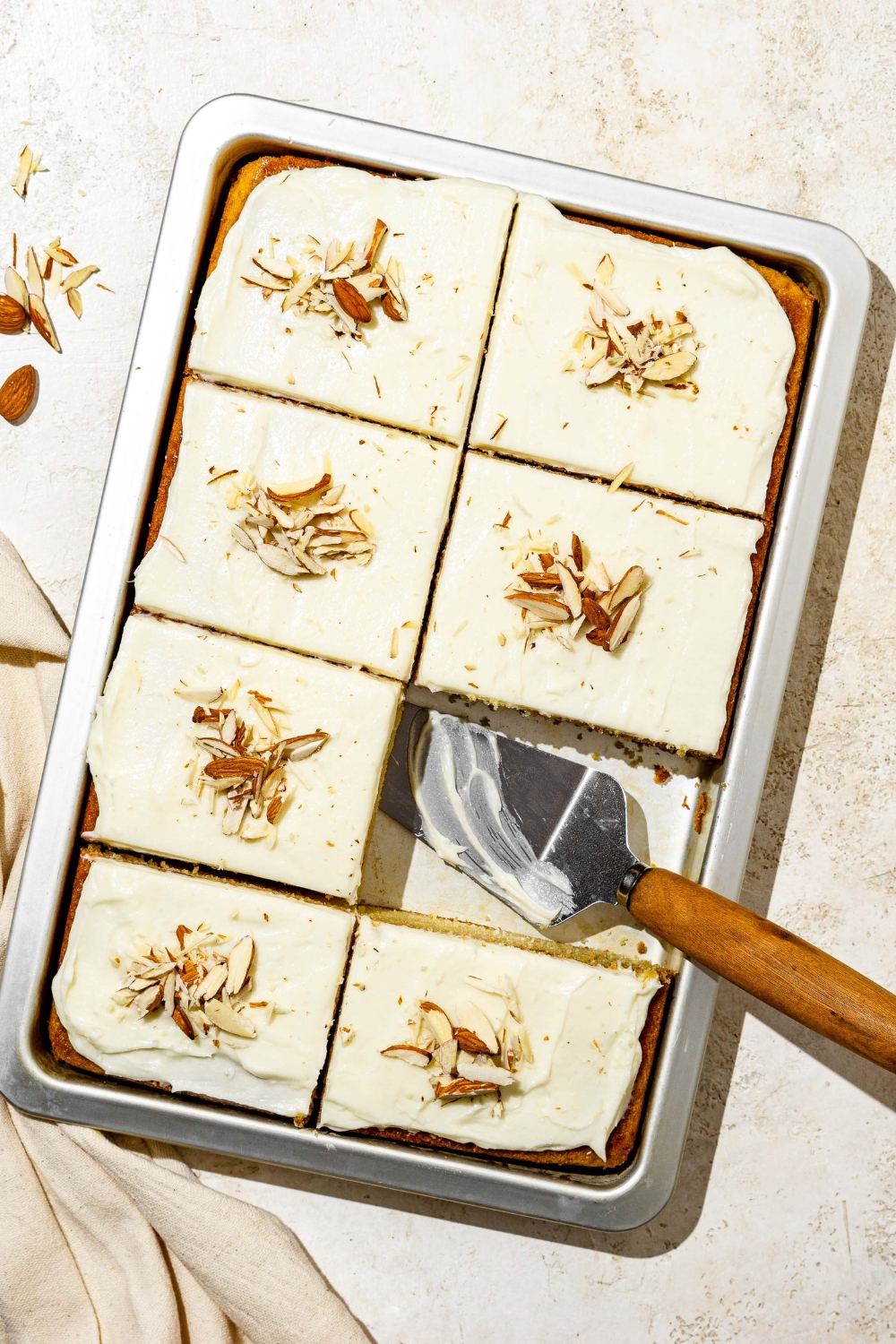 A baking pan with white Texas sheet cake topped with icing and sliced into squares. Each slice is topped with sliced almonds. A slice has been removed and there is a serving spatula.