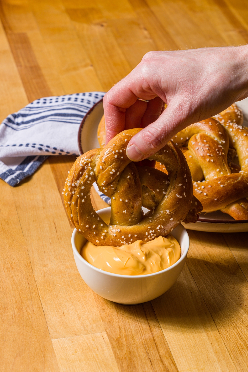 A hand dipping a copycat Auntie Anne's pretzel in a bowl of pretzel cheese. The bowl is on a wood counter with a plate of pretzels.