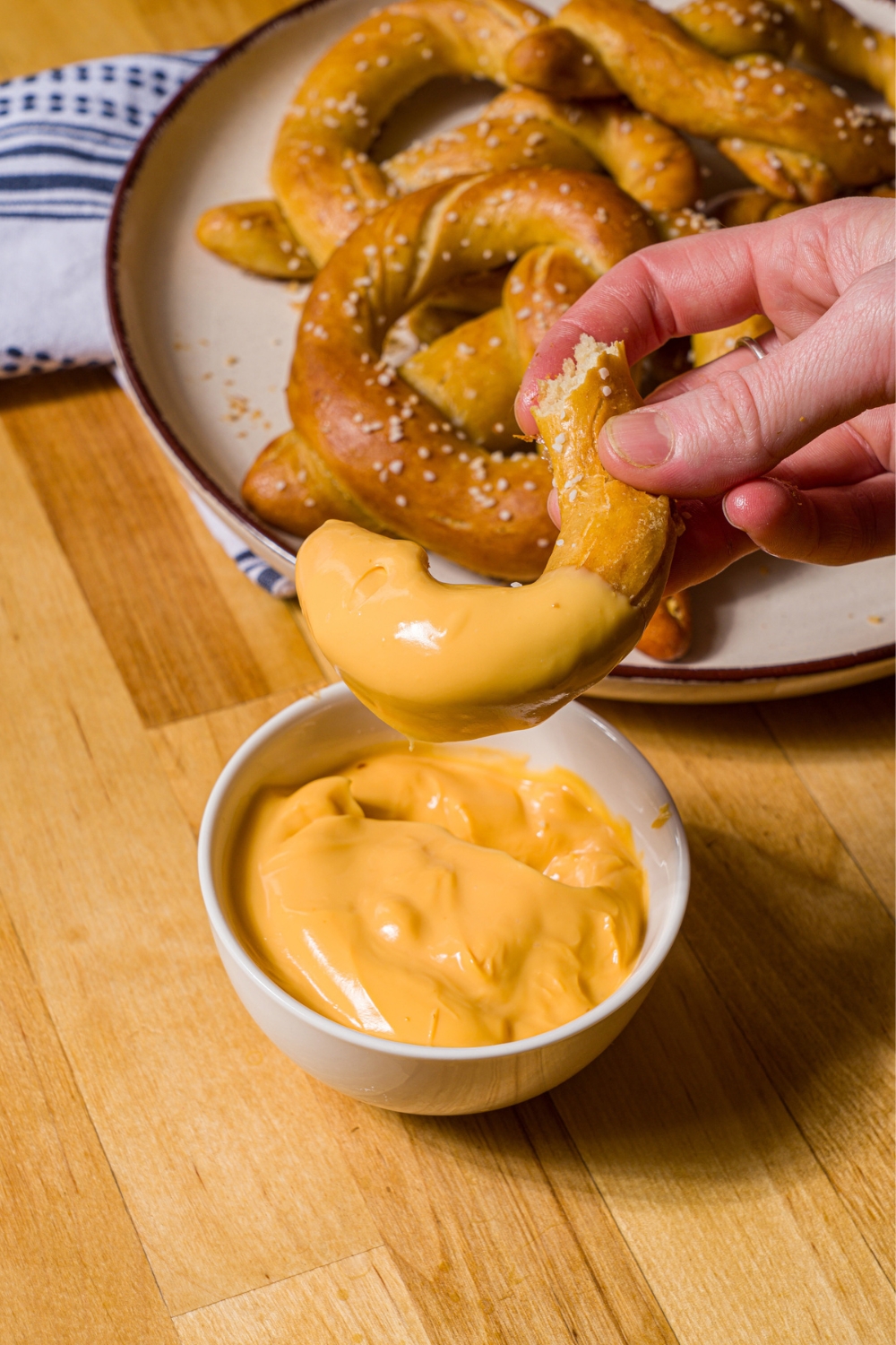 A hand dipping a piece of copycat Auntie Anne's pretzels into a bowl of pretzel cheese. The bowl is on a wood counter with a plate of pretzels and blue striped napkin.