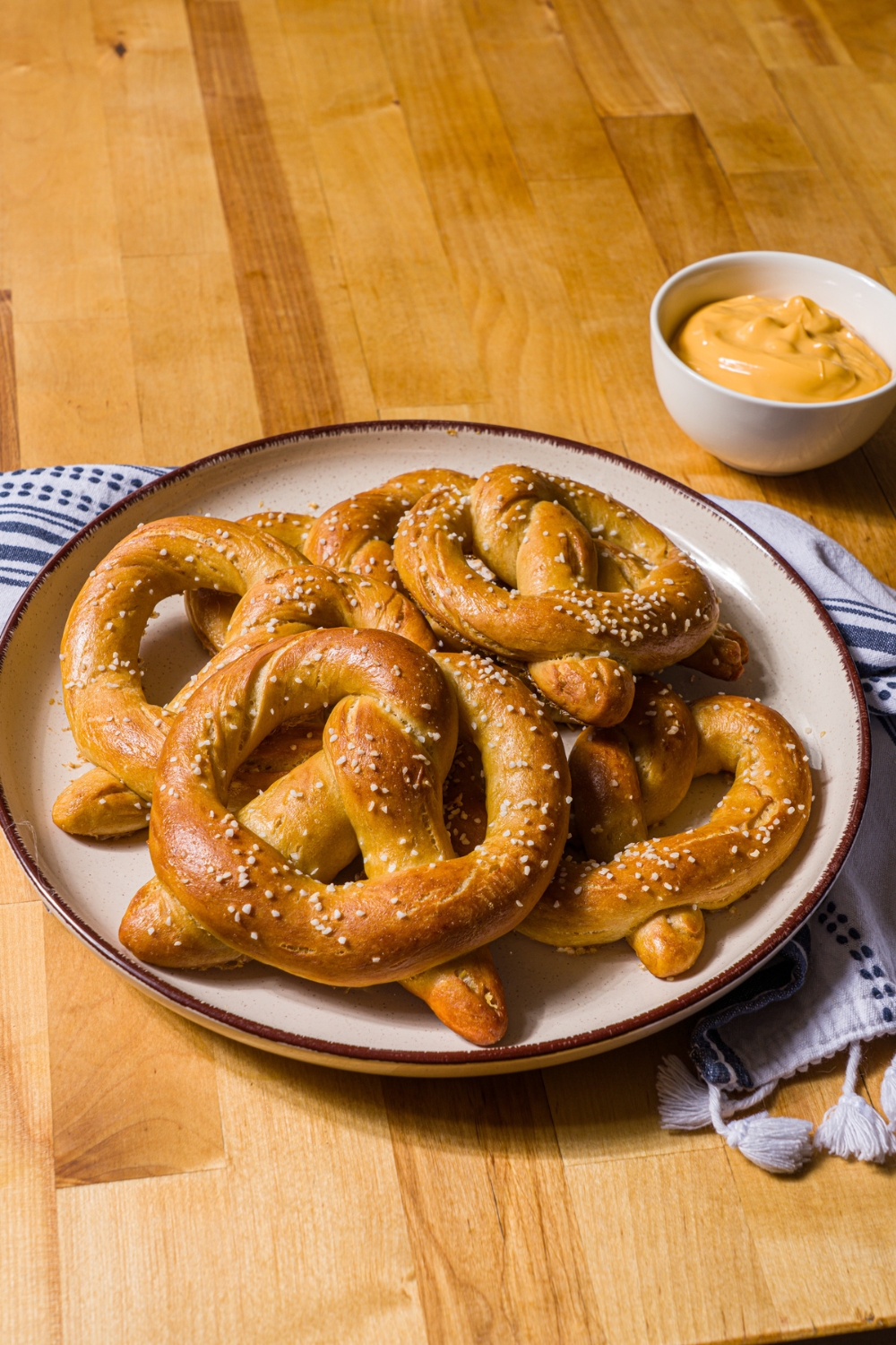 A white plate with copycat Auntie Anne's pretzels on a blue striped napkin on a wood counter with a bowl of pretzel cheese.