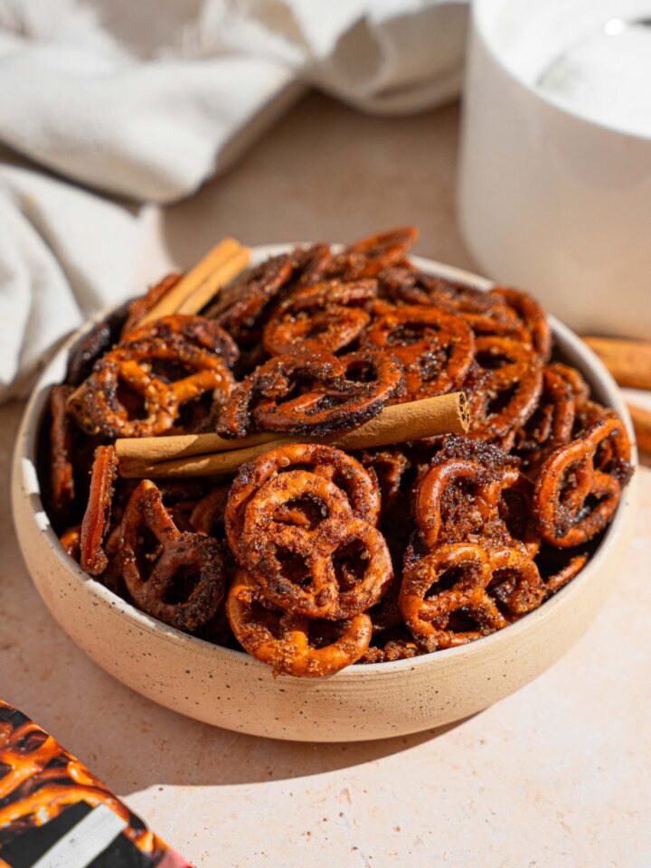 A bowl of cinnamon sugar pretzels tossed with cinnamon sticks. The bowl is on a tan counter with a white cloth napkin.