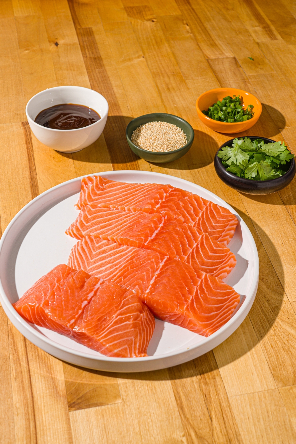 A wood counter with ingredients to make baked teriyaki salmon including uncooked salmon fillets, teriyaki sauce, sesame seeds, green onion, and cilantro.