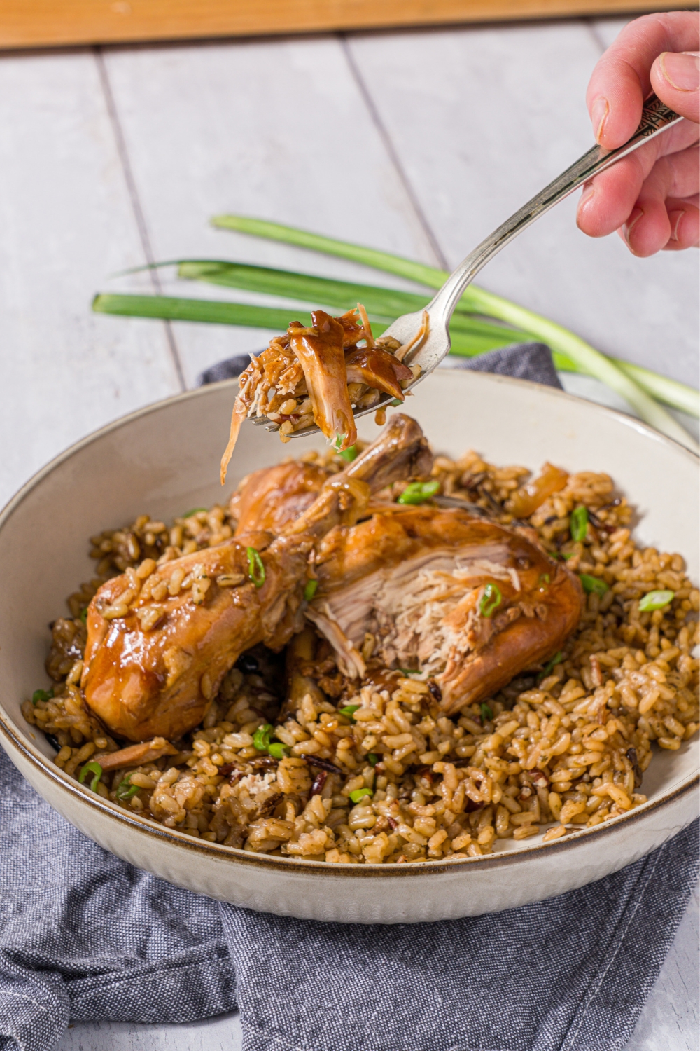 A bowl of slow cooker chicken adobo served over brown rice garnished with sliced green onion. There is a fork with a bite of chicken held over the bowl. The bowl is on a wood counter with a blue cloth napkin.