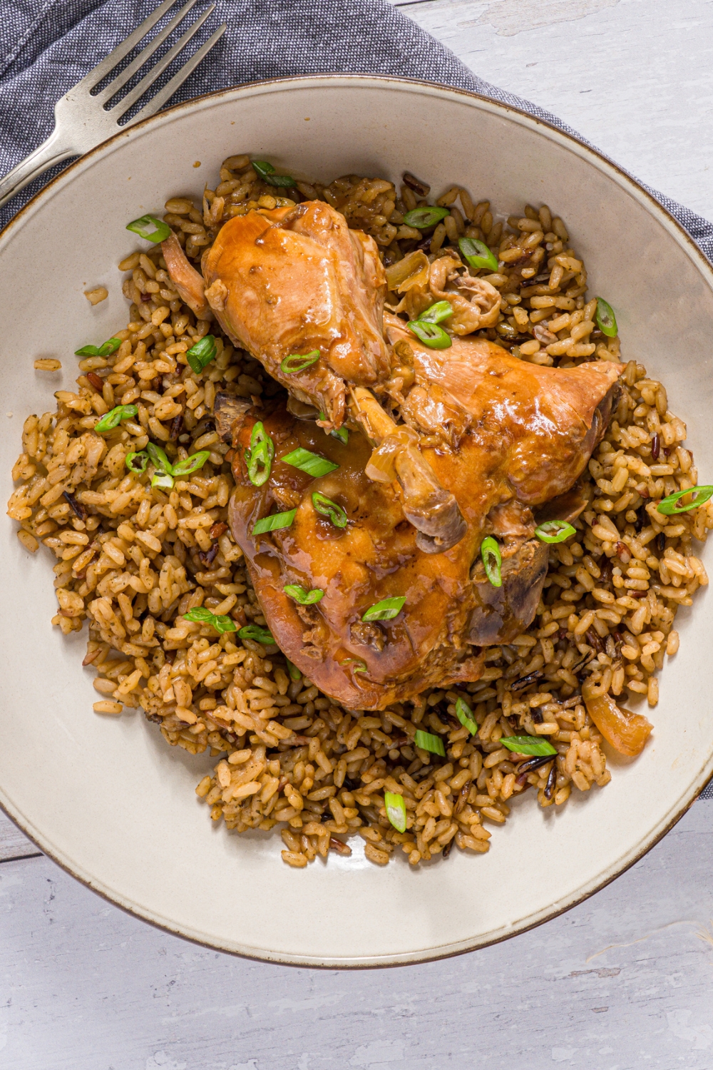 A bowl of slow cooker chicken adobo served over brown rice garnished wtih sliced green onion. The bowl is on a wood counter with a blue cloth napkin.