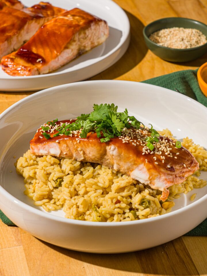 A white bowl with baked teriyaki salmon garnished with sesame seeds, sliced green onions, and cilantro over fried rice. The bowl is on a wood counter with a plate of baked salmon.