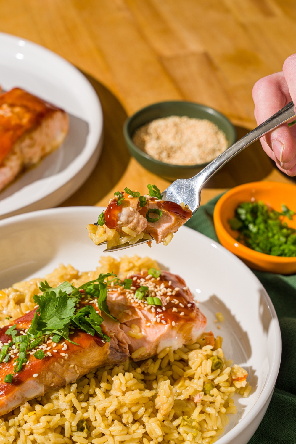 A fork with a bite of baked teriyaki salmon over a bowl of salmon garnished with sesame seeds, sliced green onion, and cilantro. The salmon is served over fried rice. The bowl is on a wood counter with bowls of garnishes.