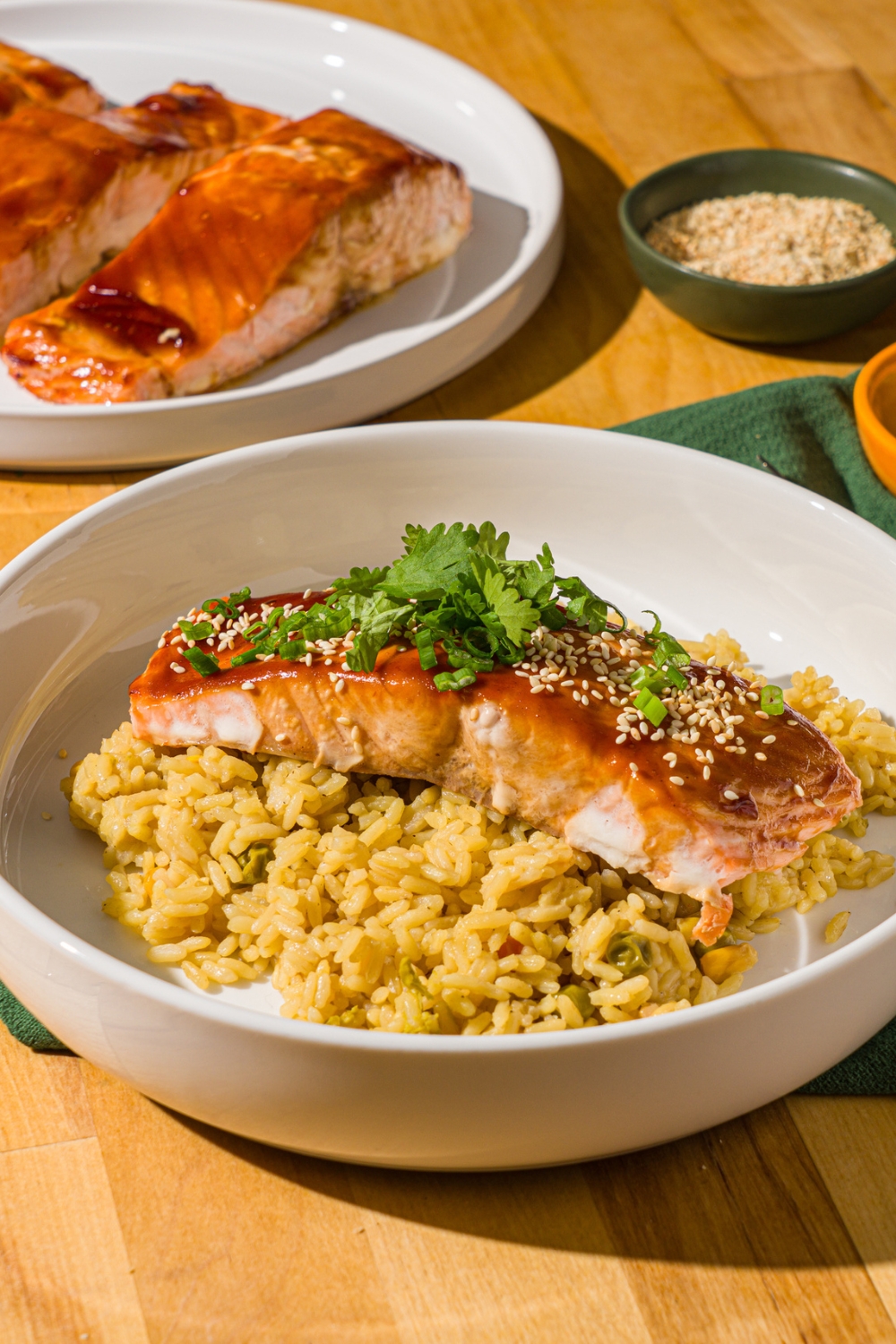 A white bowl with baked teriyaki salmon garnished with sesame seeds, sliced green onions, and cilantro over fried rice. The bowl is on a wood counter with a plate of baked salmon.
