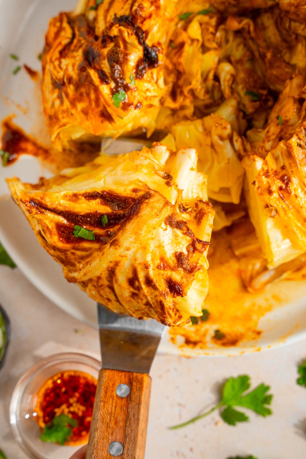 A plate with a baked cabbage boil with cajun seasoned butter. There is a spatula taking a piece of cabbage from the plate. The plate is on a tan counter with a small bowl of seasoned butter and fresh parsley.