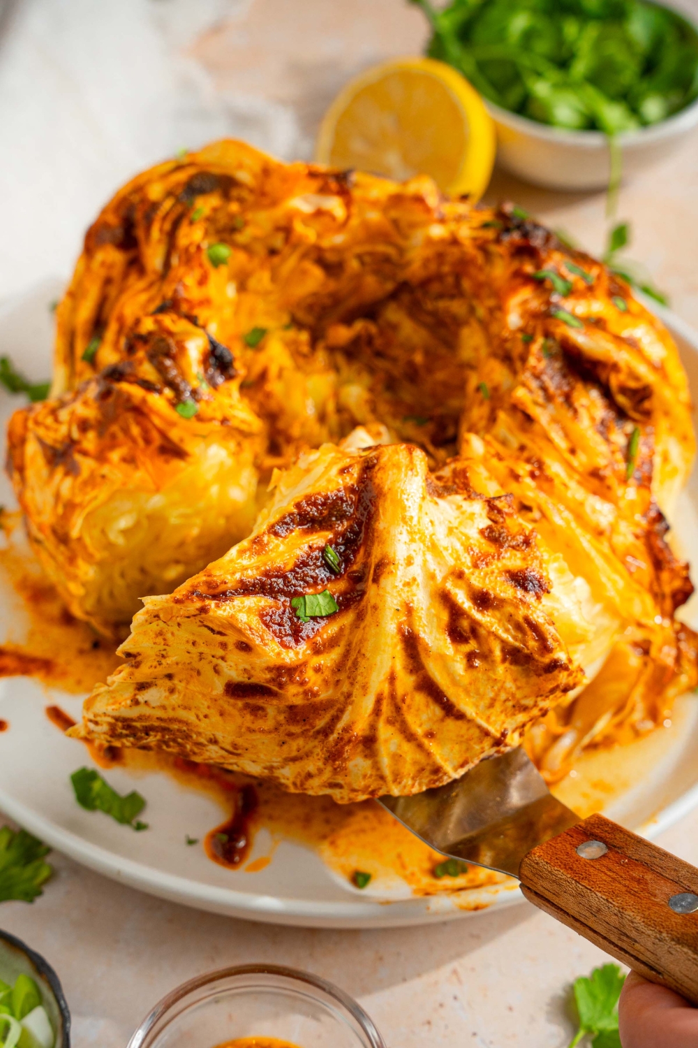 A plate with a baked cabbage boil with cajun seasoned butter. There is a spatula taking a piece of cabbage from the plate. The plate is on a tan counter with lemon and fresh parsley.