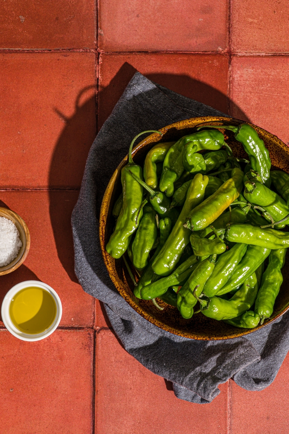 A bowl of shishito peppers with a bowl of flaky salt and small bowl of oil on a red tiled counter with a blue cloth napkin.