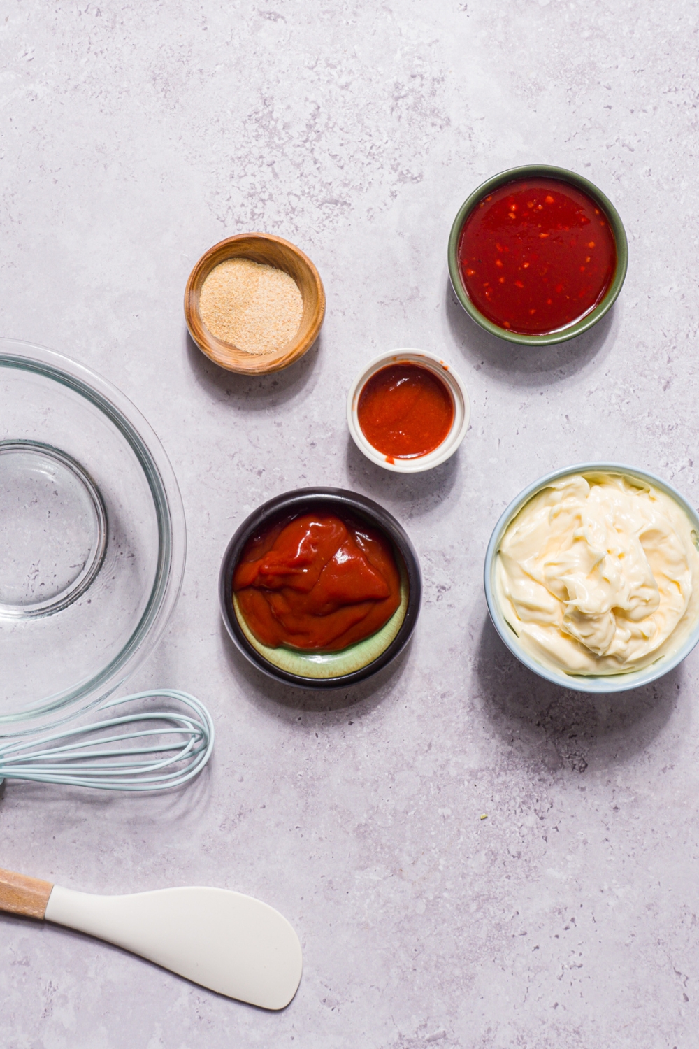 A stone counter with several bowls in various sizes containing ingredients to make boom boom sauce including mayo, ketchup, sriracha, sweet chili sauce, and seasonings.