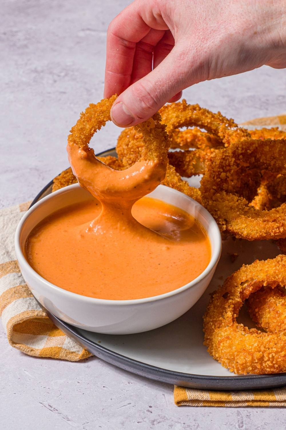 A plate of fried onion rings with a bowl of boom boom sauce. A hand is dipping an onion ring into the sauce. The plate is on a stone counter with a yellow striped napkin.
