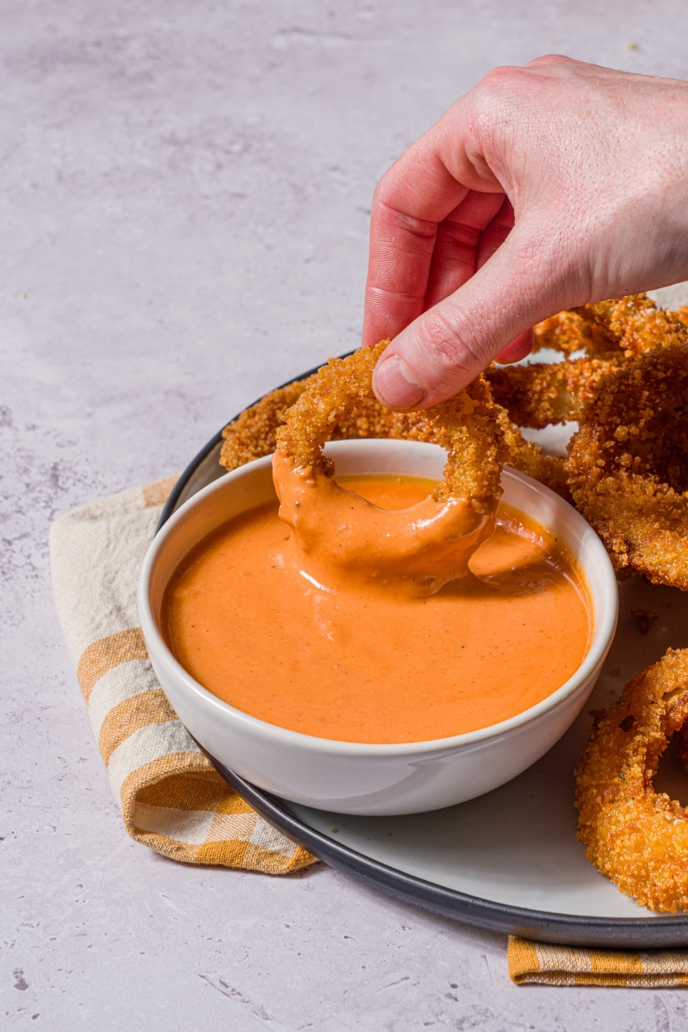 A plate of fried onion rings with a bowl of boom boom sauce. A hand is dipping an onion ring into the sauce. The plate is on a stone counter with a yellow striped napkin.