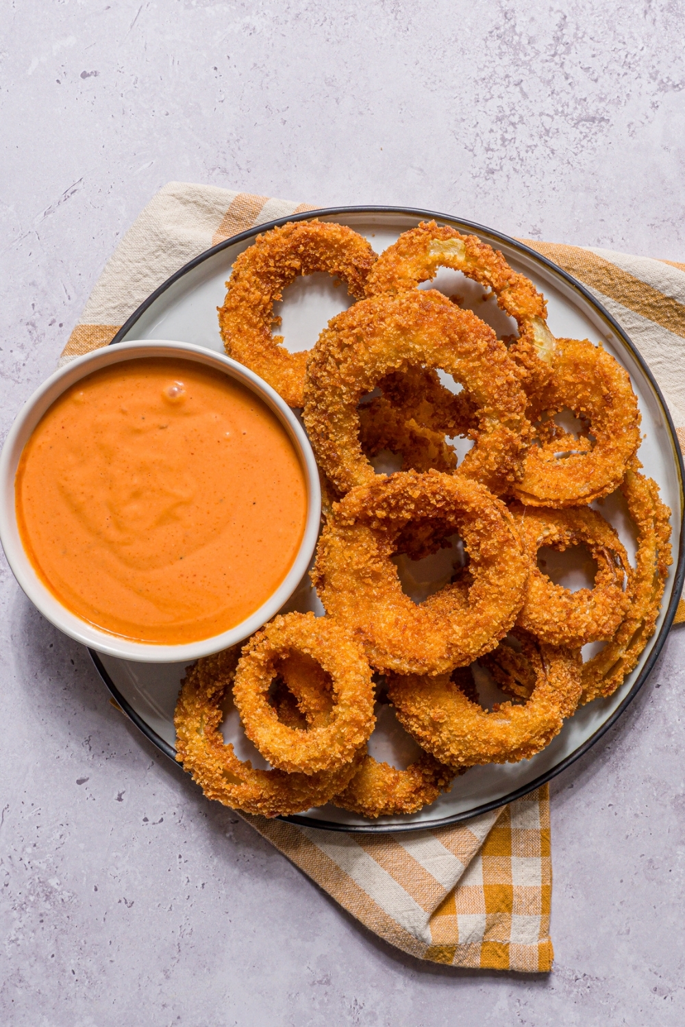 A plate of fried onion rings with a bowl of boom boom sauce. The plate is on a stone counter with a yellow striped napkin.