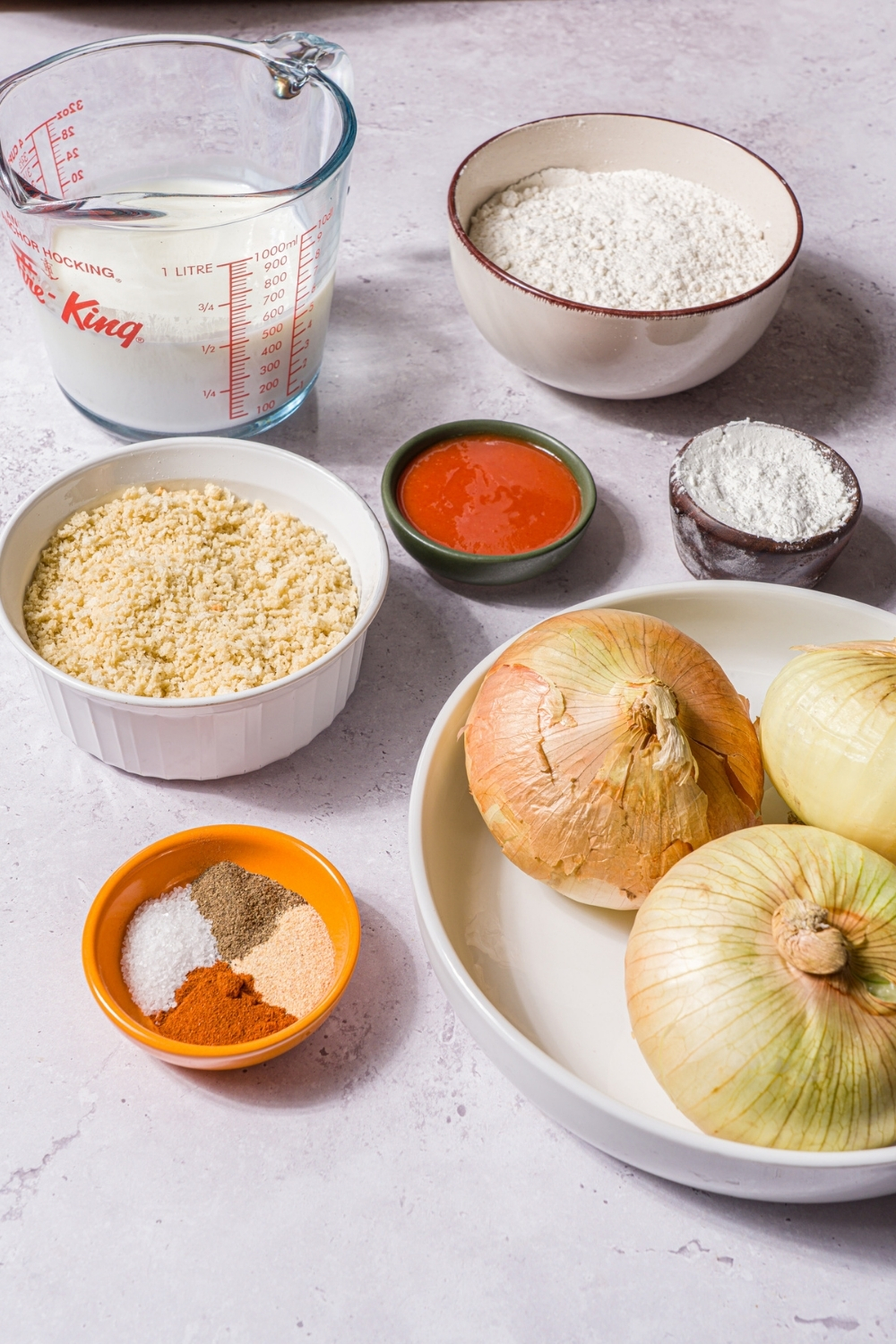 A stone counter with ingredients to make onion rings including onions, breading, hot sauce, buttermilk, flour, and seasonings.