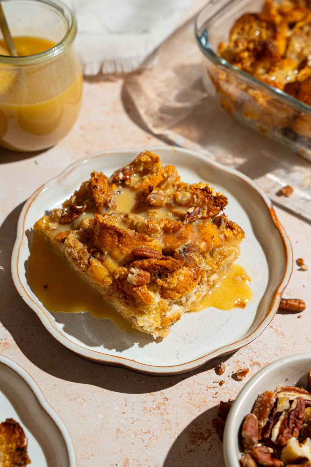 A white plate with a slice of bread pudding topped with rum sauce. The plate is on a tan counter with a baking dish of bread pudding and jar of rum sauce.