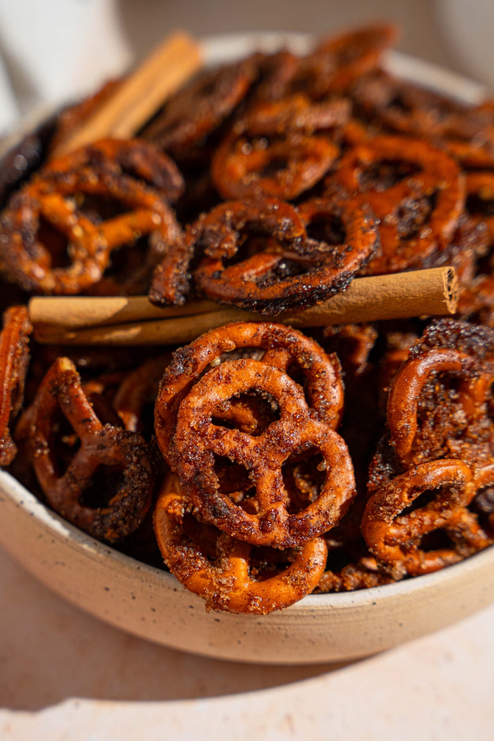 A bowl of cinnamon sugar pretzels tossed with cinnamon sticks. The bowl is on a tan counter.