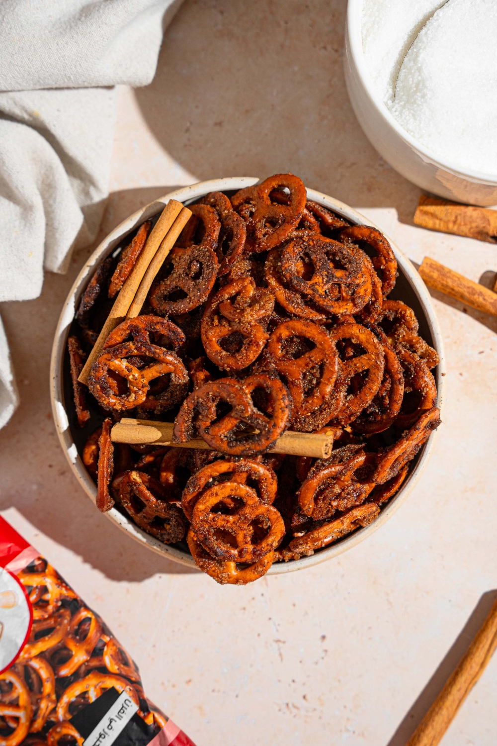 A bowl of cinnamon sugar pretzels tossed with cinnamon sticks. The bowl is on a tan counter with a white cloth napkin.