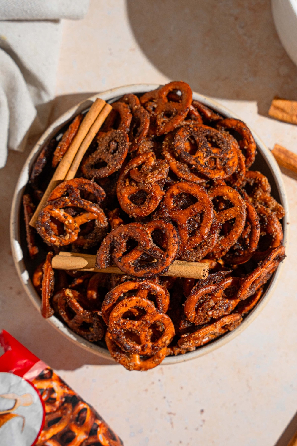 A bowl of cinnamon sugar pretzels tossed with cinnamon sticks. The bowl is on a tan counter with a white cloth napkin.