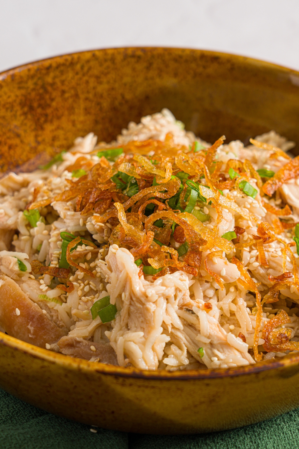 A brown ceramic bowl with viral Costco chicken, scallion, and rice topped with sliced scallions and crispy fried onions. The bowl is on a white counter with a green cloth napkin.