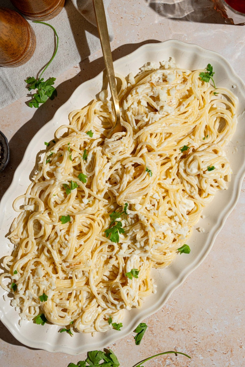 A white platter with Mexican white spaghetti garnished with fresh parsley. The spaghetti is tossed with crumbled queso fresco. There is a fork in the spaghetti. The platter is on a tan counter.