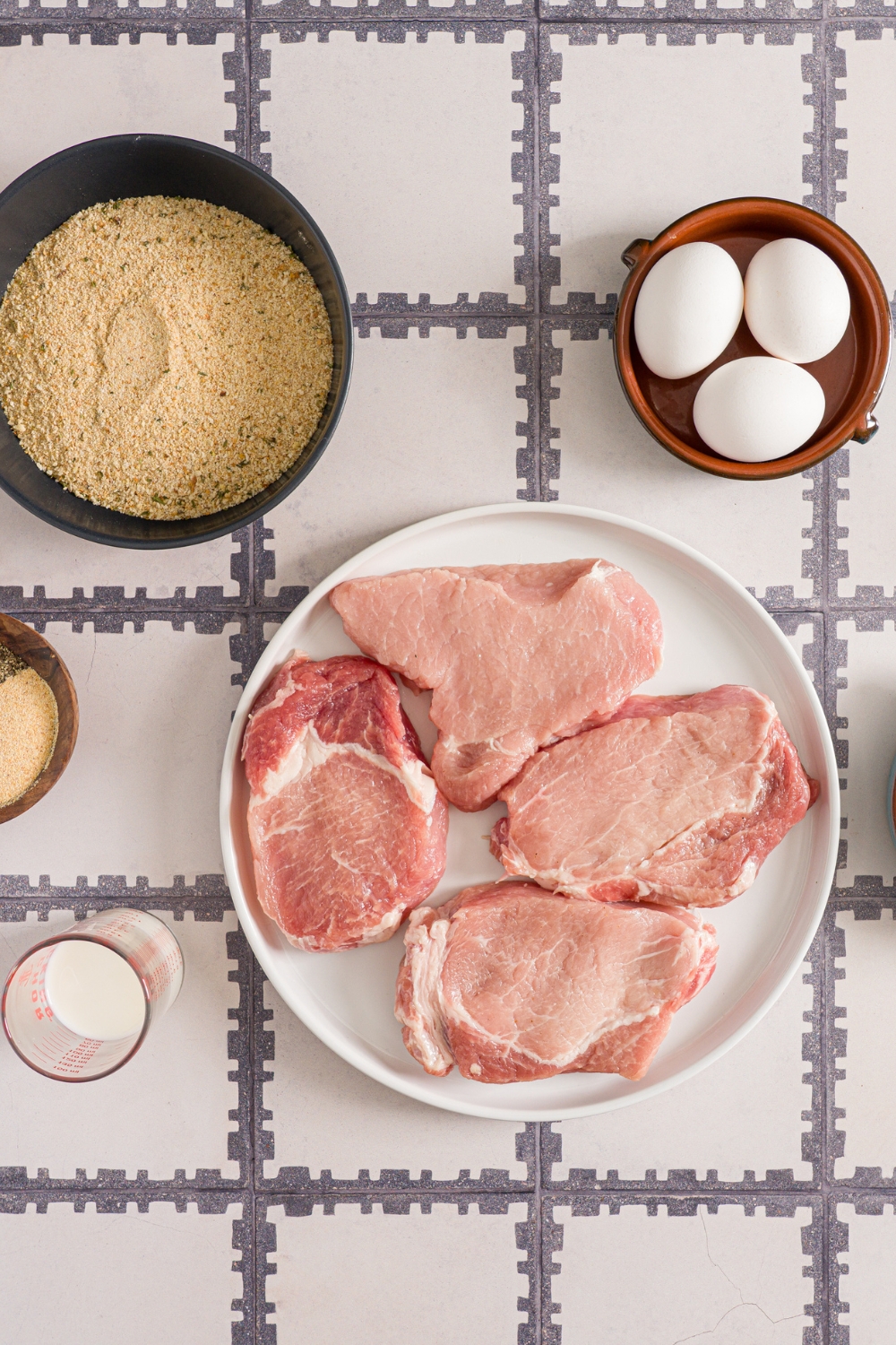 Several bowls in various sizes containing ingredients to make crispy breaded pork chops including pork cutlets, eggs, panko breading, and milk.
