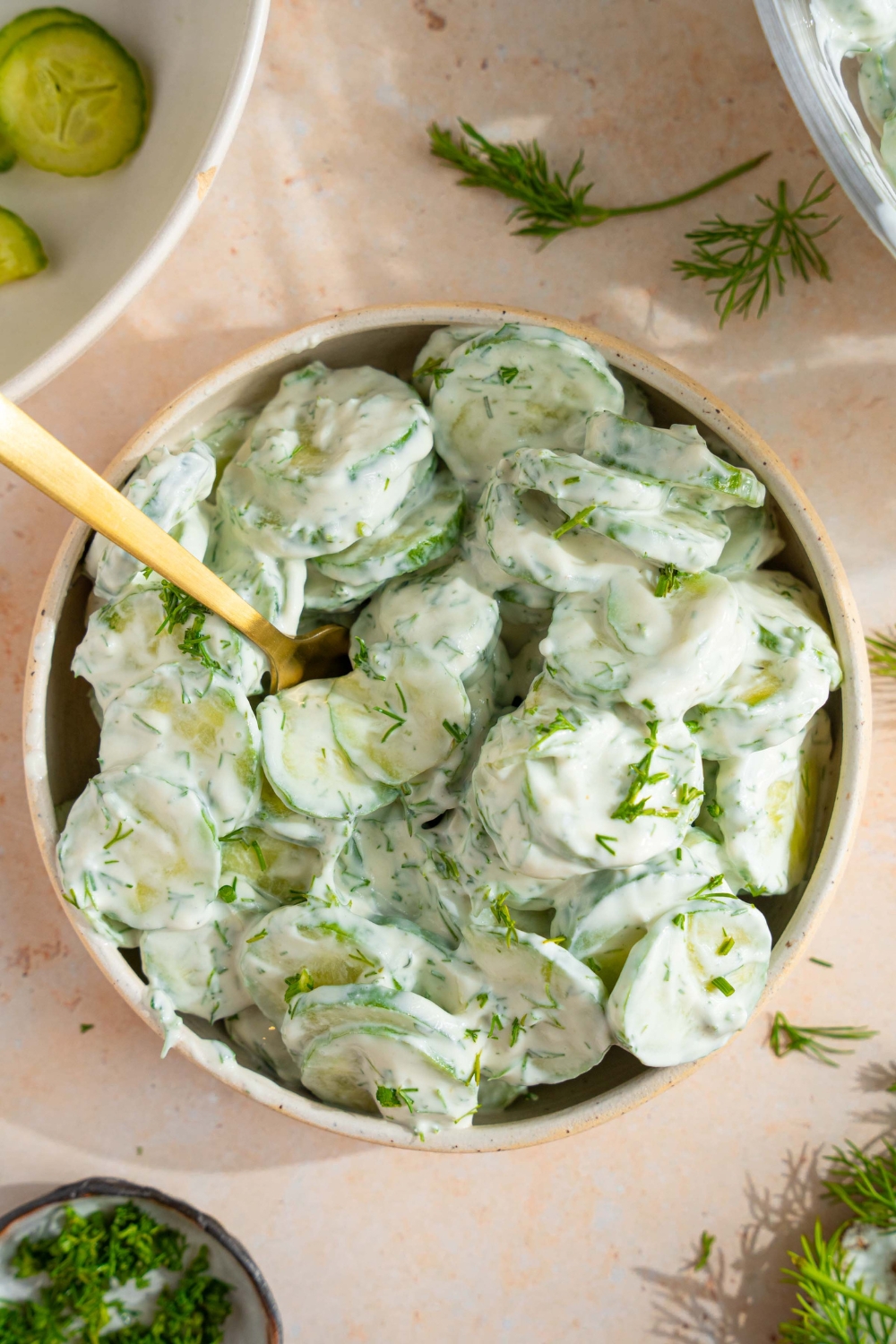A bowl of cucumber dill salad tossed with a yogurt and sour cream dressing with fresh dill. There is a fork in the bowl. The bowl is on a tan counter with sprigs of dill and bowl of sliced cucumber.