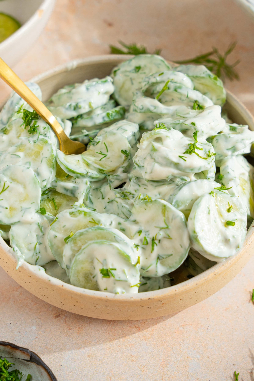A bowl of cucumber dill salad tossed with a yogurt and sour cream dressing with fresh dill. There is a fork in the bowl. The bowl is on a tan counter with sprigs of dill and bowl of sliced cucumber.