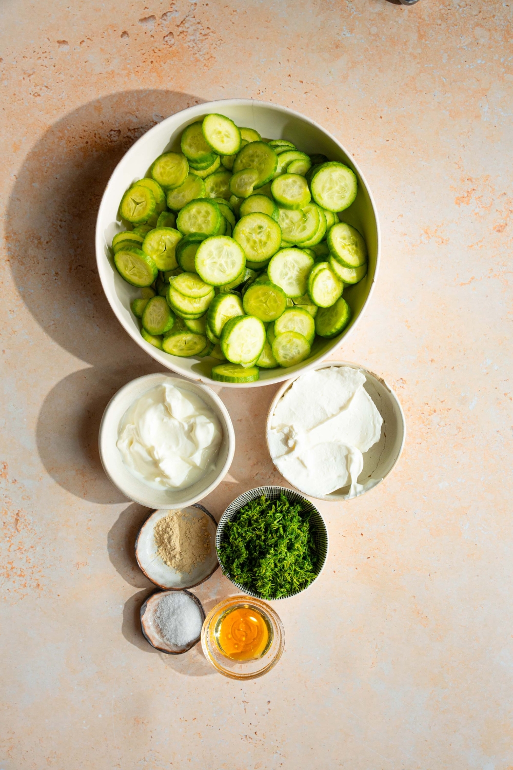 An overhead shot of several bowls in various sizes containing ingredients to make cucumber dill salad including sliced cucumber, sour cream, greek yogurt, fresh dill, honey, and seasonings.