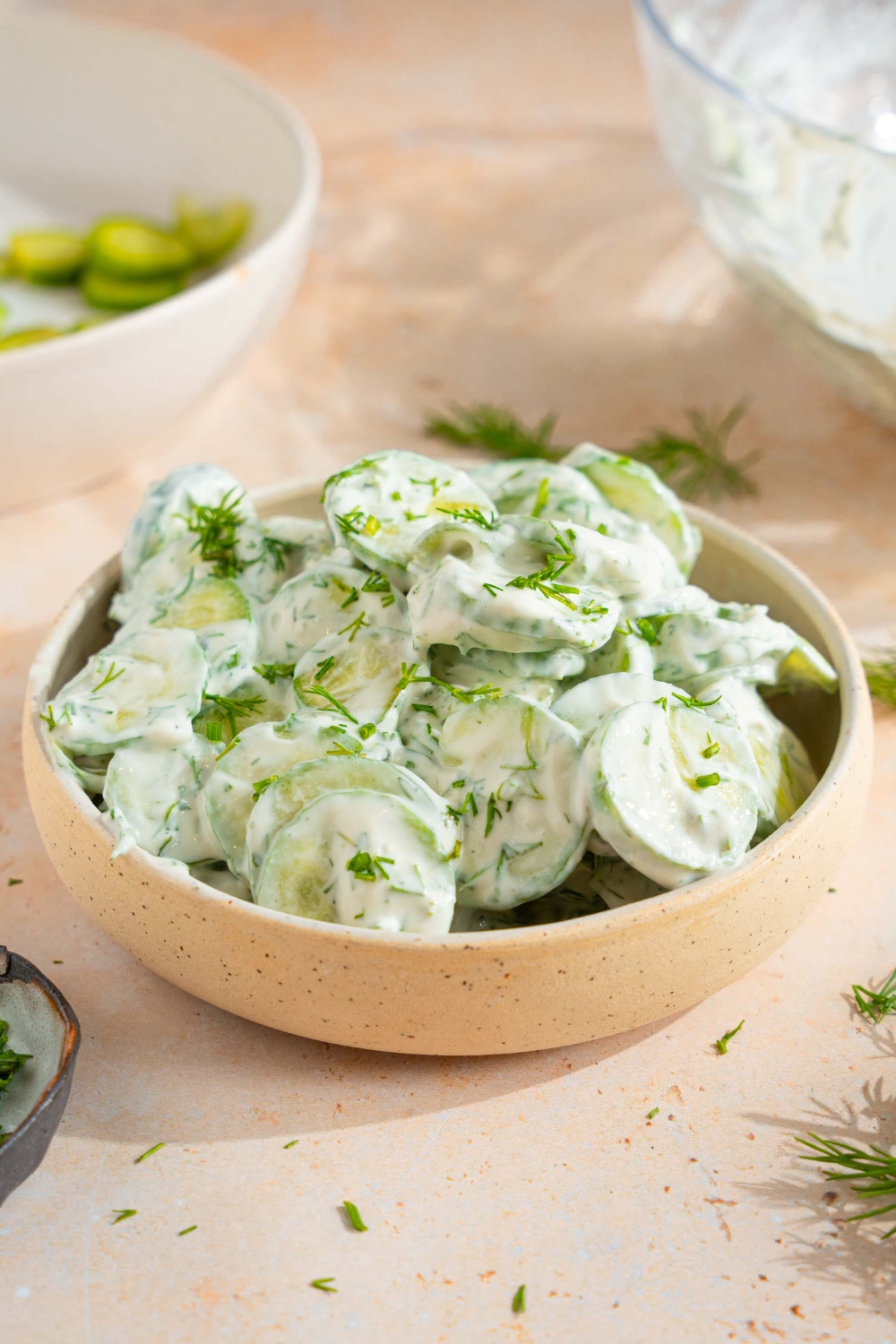 A bowl of cucumber dill salad tossed with a yogurt and sour cream dressing with fresh dill. The bowl is on a tan counter with sprigs of dill and bowl of sliced cucumber.