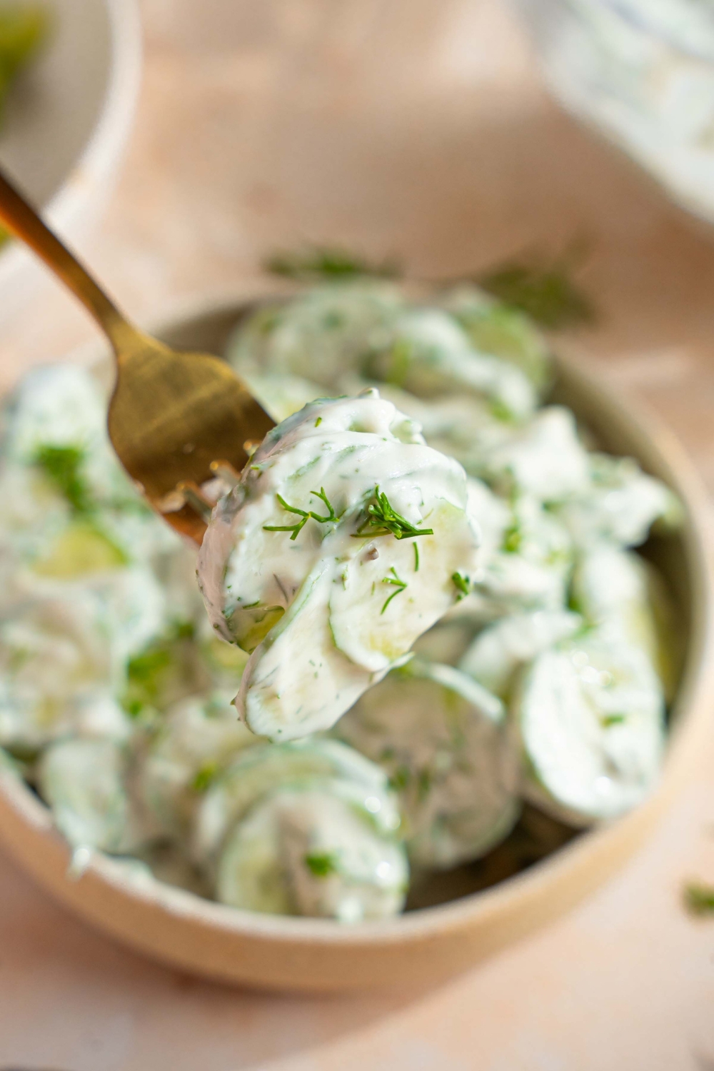 A close up of a fork with a bite of cucumber dill salad garnished with fresh dill. There is a bowl of cucumber dill salad on a tan counter blurred in the background.