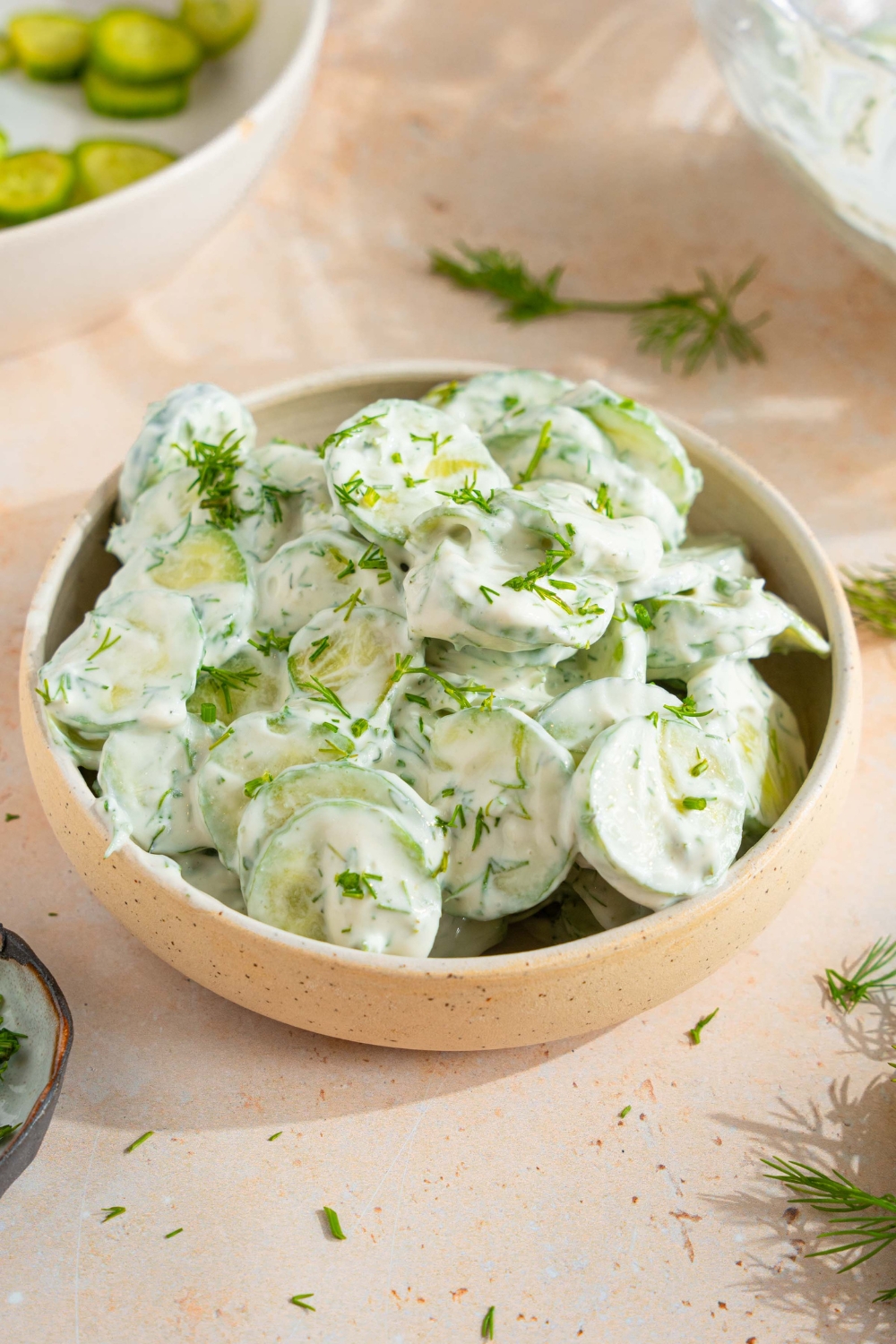 A bowl of cucumber dill salad tossed with a yogurt and sour cream dressing with fresh dill. The bowl is on a tan counter with sprigs of dill and bowl of sliced cucumber.