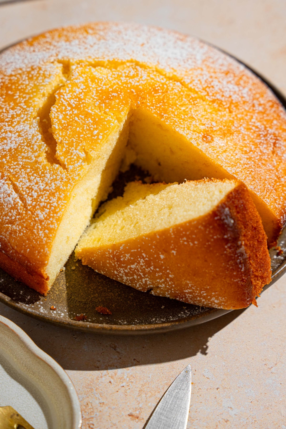 A green plate with a whole French butter cake dusted with powdered sugar. There is a slice removed from the cake on the plate. The plate is on a tan counter.