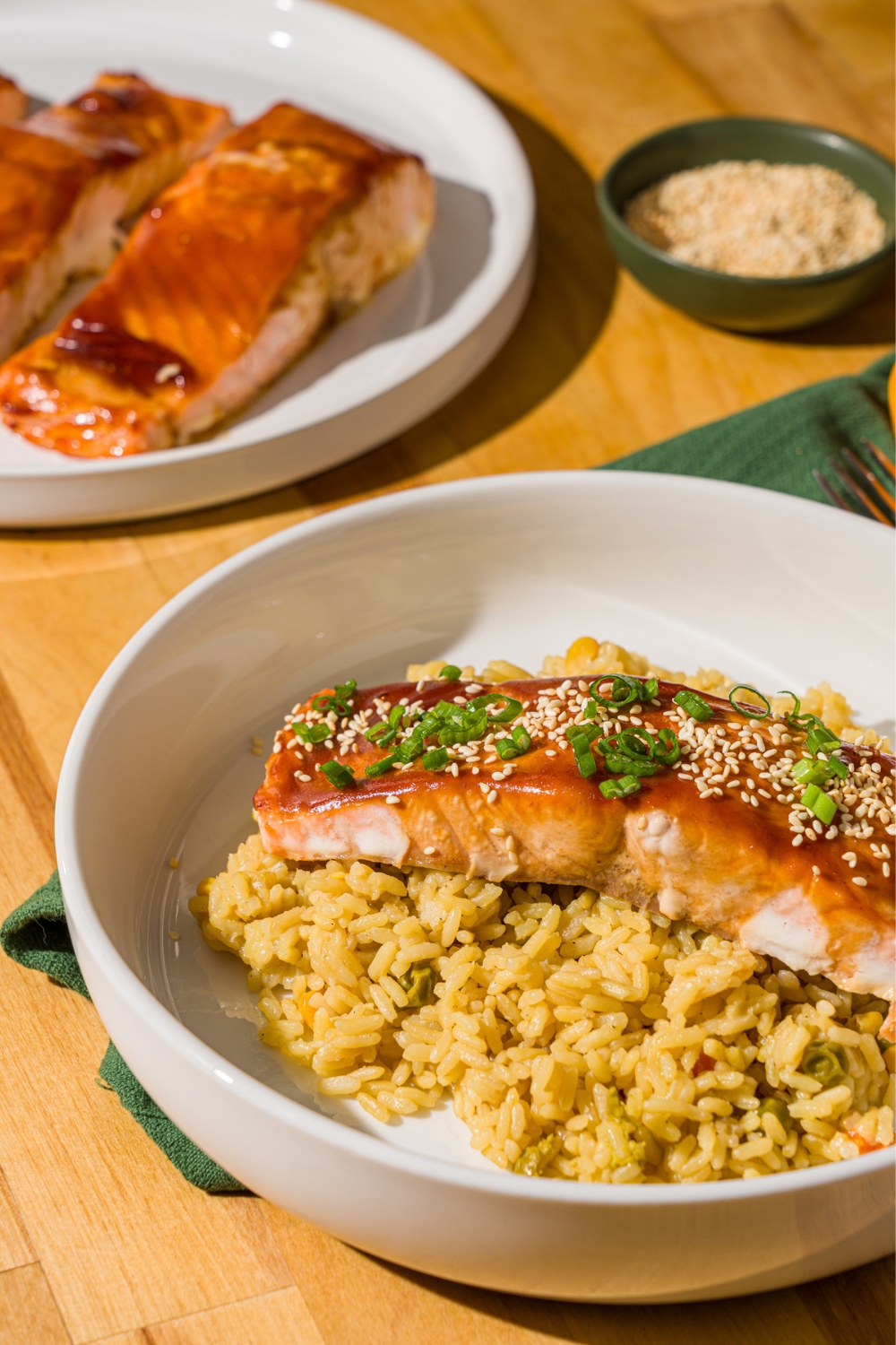 A white bowl with baked teriyaki salmon garnished with sesame seeds, sliced green onions, and cilantro over fried rice. The bowl is on a wood counter with a plate of baked salmon.