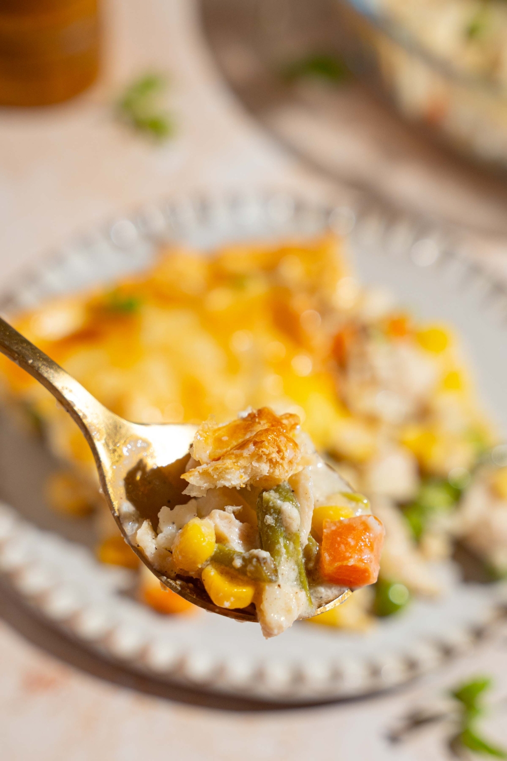 A close up of a spoon with a bite of chicken pot pie. There is a plate with a slice of pot pie on a tan counter blurred in the background.