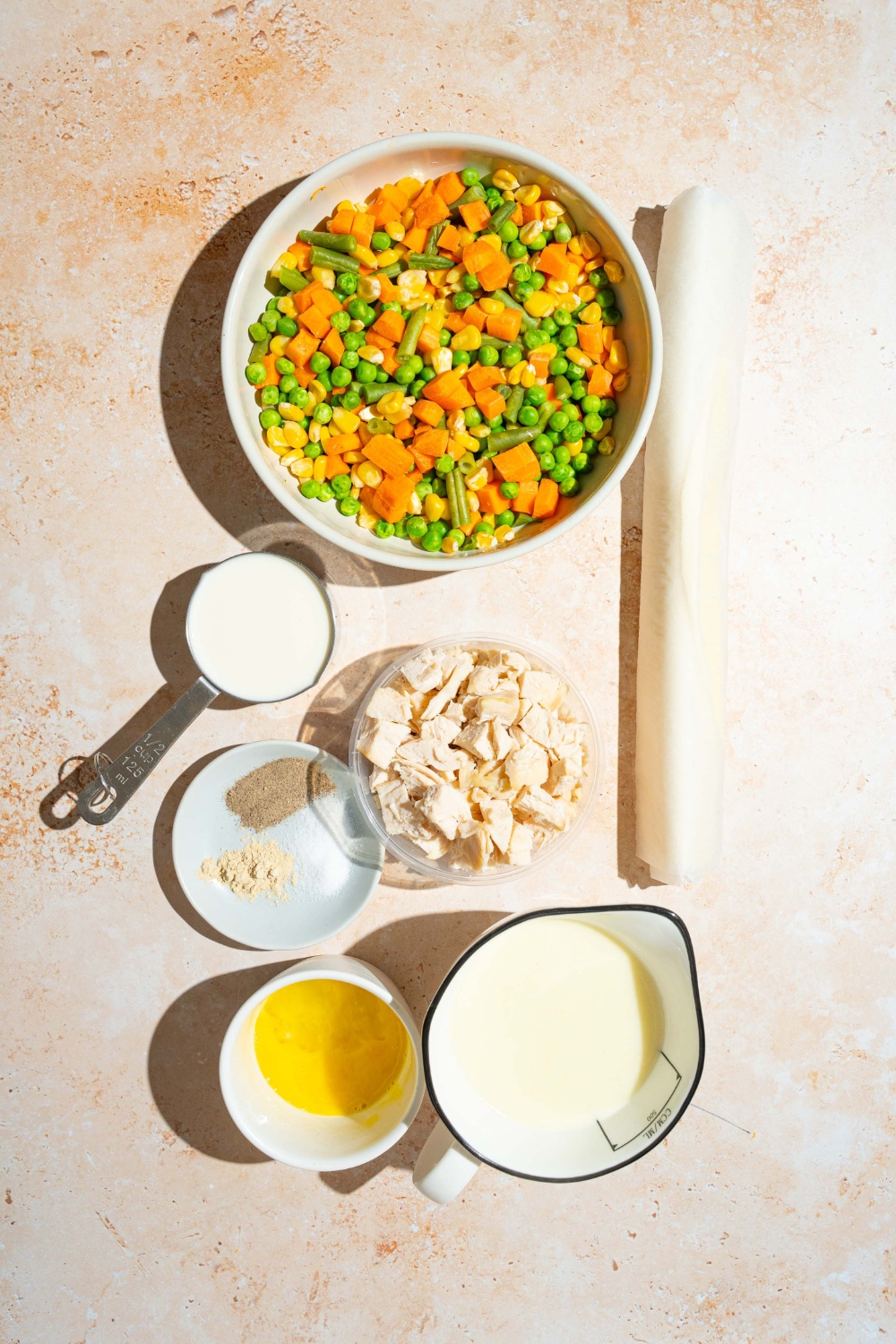 An overhead shot of several bowls in various sizes containing ingredients to make chicken pot pie including frozen mixed vegetables, cream of chicken soup, cubed chicken, milk, prepackaged pie crust roll, and seasonings.