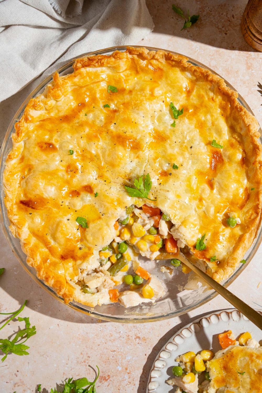 A whole chicken pot pie garnished with fresh parsley with a slice of pie removed. A spoon is in the pie dish. The dish is on a tan counter with a plate with a slice of pot pie.