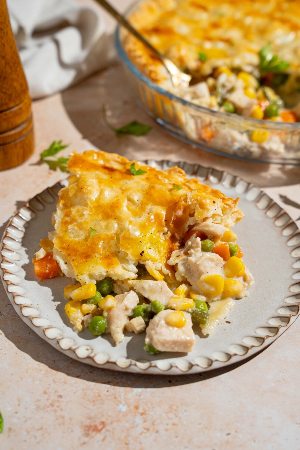 A plate with a slice of chicken pot pie garnished with fresh parsley. The plate is on a tan counter with a pie dish of pot pie.
